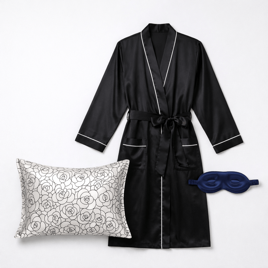 Black silk robe with white piping, floral pillow, and blue sleep mask on a light gray background