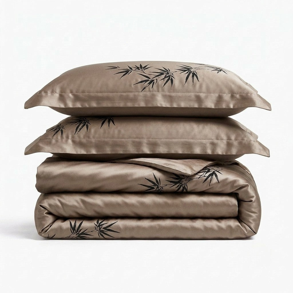 A stack of two pillows and a folded duvet cover in a warm taupe-brown silk fabric, both printed with a black bamboo leaf pattern, set against a plain white background.
