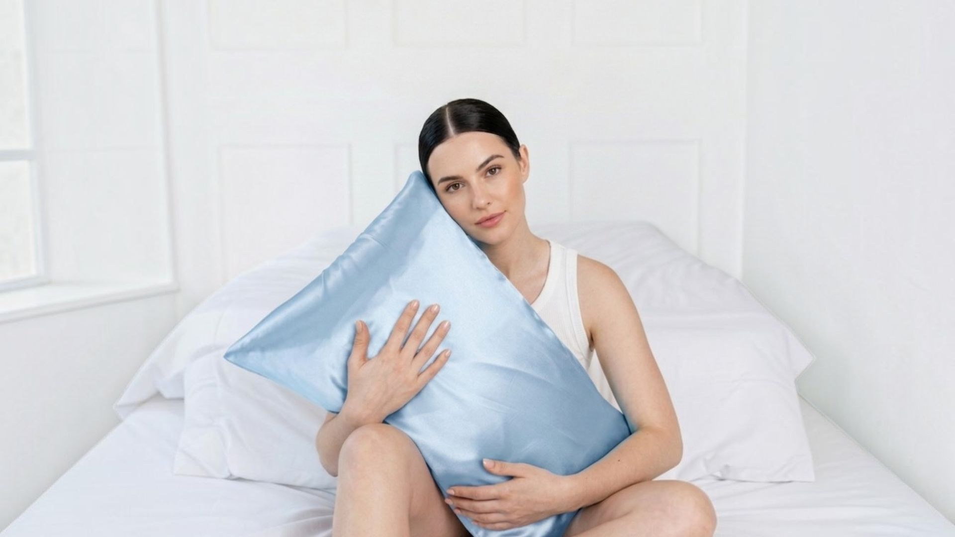 A woman sits on a white bed holding a light blue silk pillowcase.